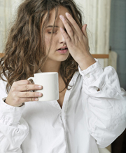 Woman dressed in white cotton shift holds her head and coffee cup